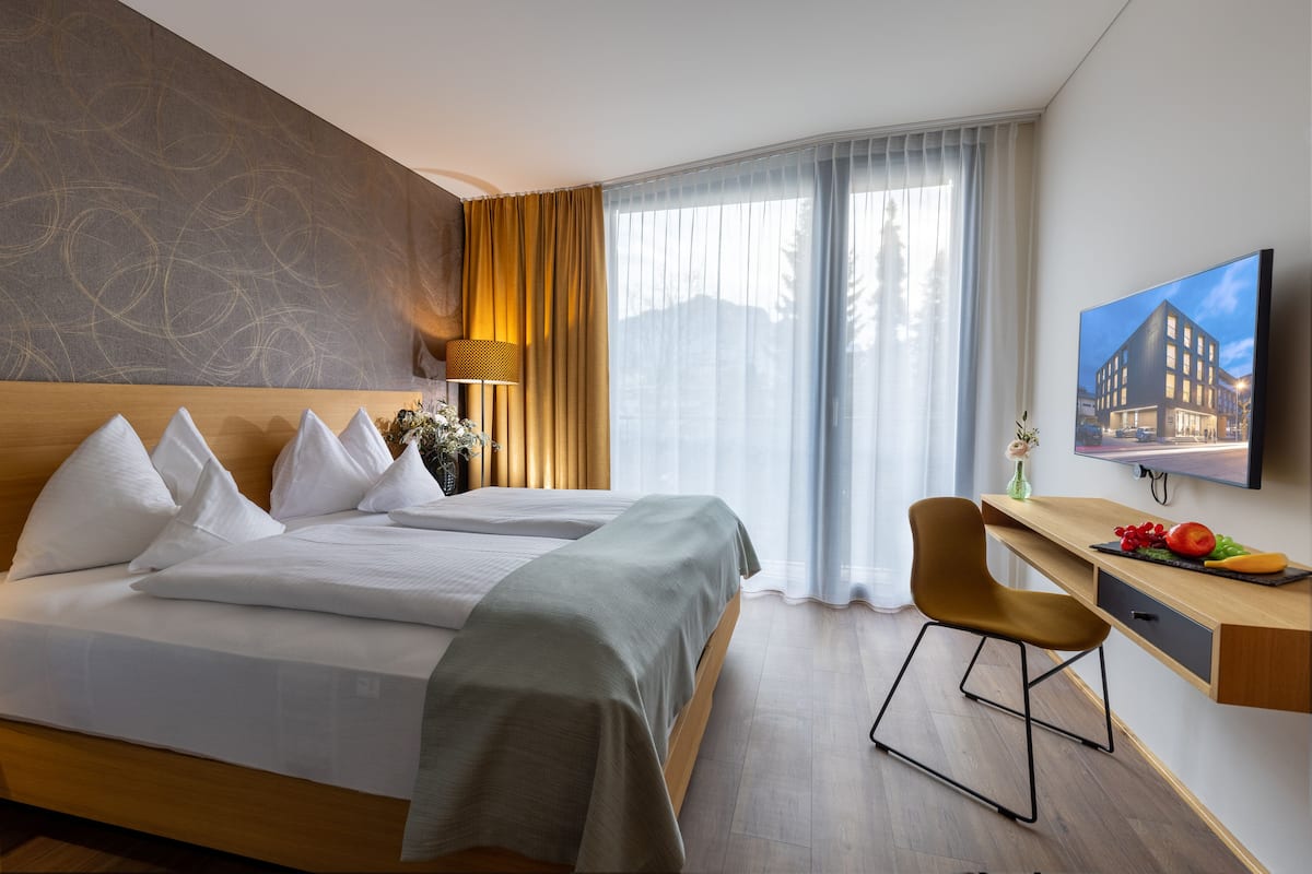 Ein Hotelzimmer mit zwei Betten, einem Schreibtisch mit Stuhl, einem Fernseher und einem großen Fenster mit Vorhängen.