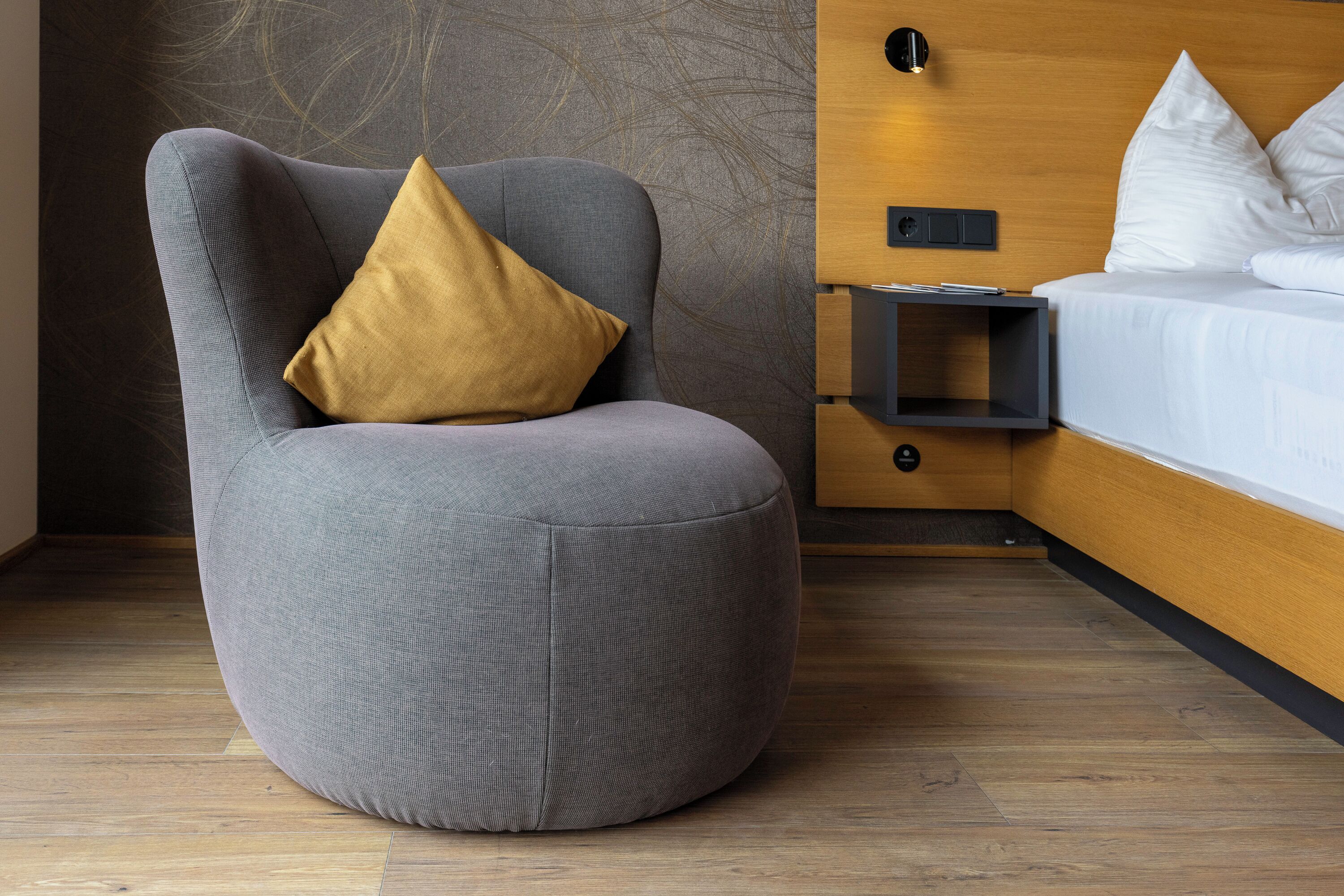 A modern room with a grey armchair, a wooden nightstand, and a bed with white bedding.