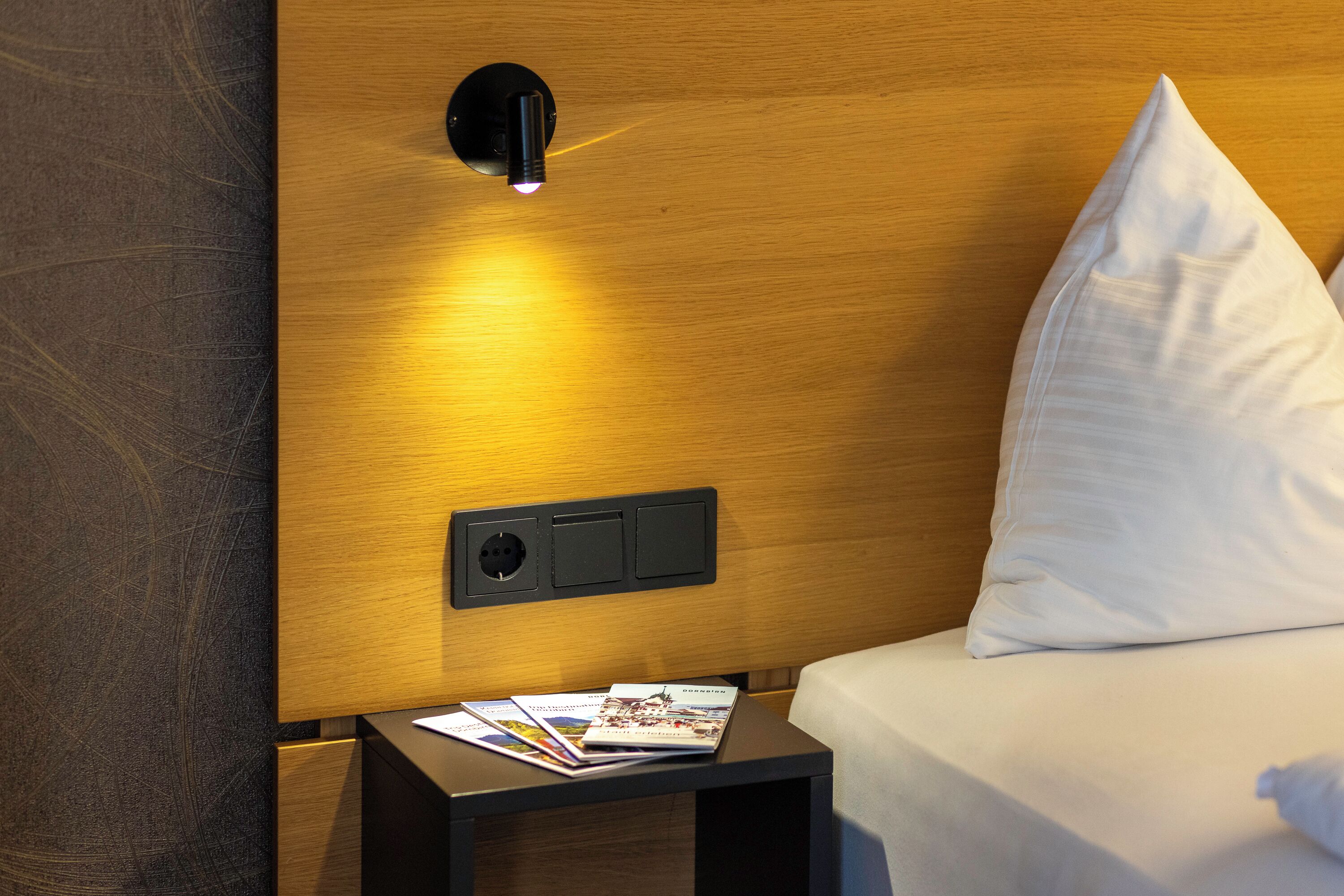 Una cama bien hecha con un almohadón blanco, una mesita de noche con folletos y una lámpara fijada a la pared.