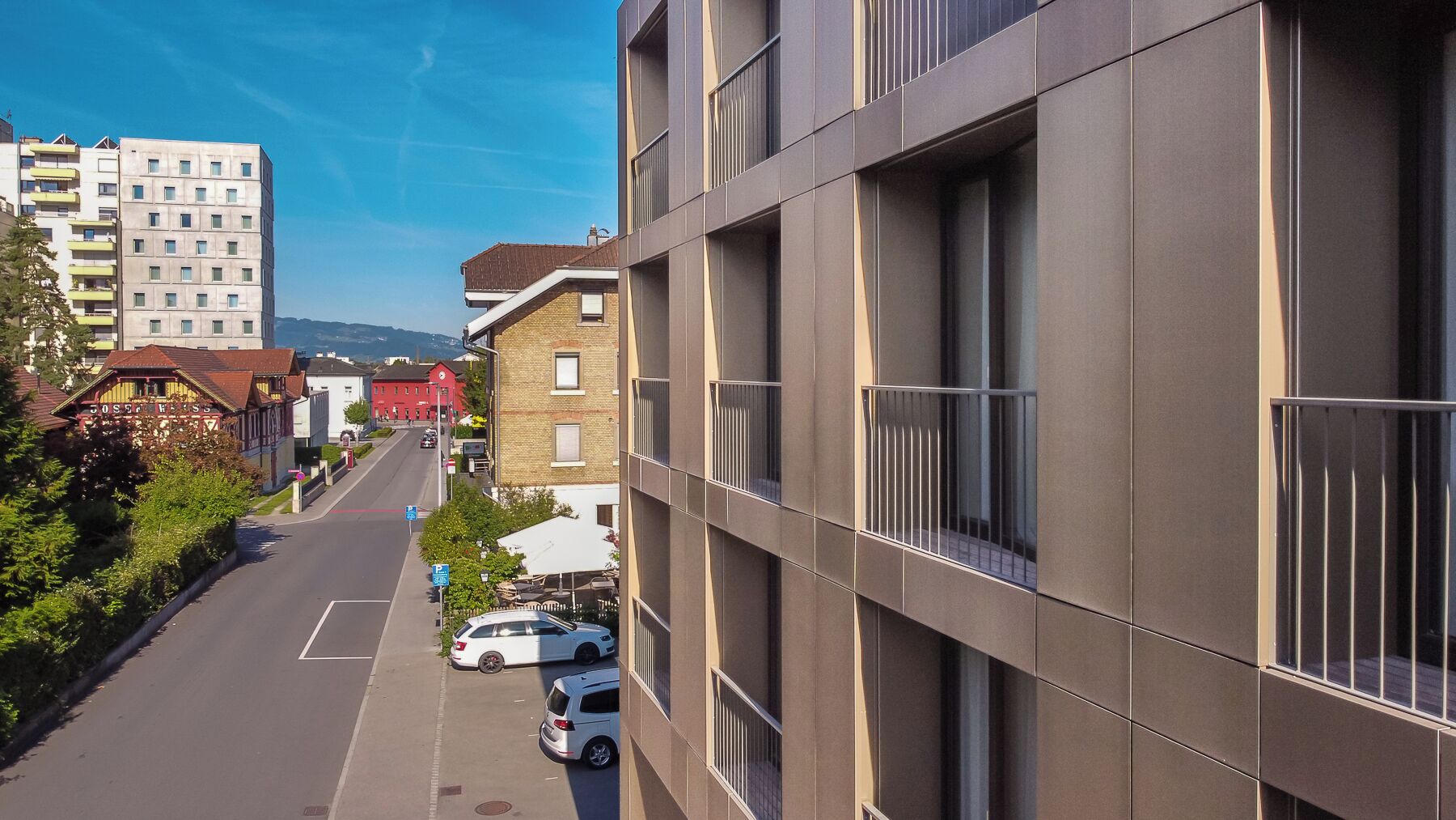 A modern building with a flat facade, large windows, and a street view with parked cars and a clear sky.