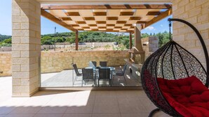 Une terrasse extérieure couverte, avec une table et des chaises, un fauteuil suspendu et un mur en pierre.