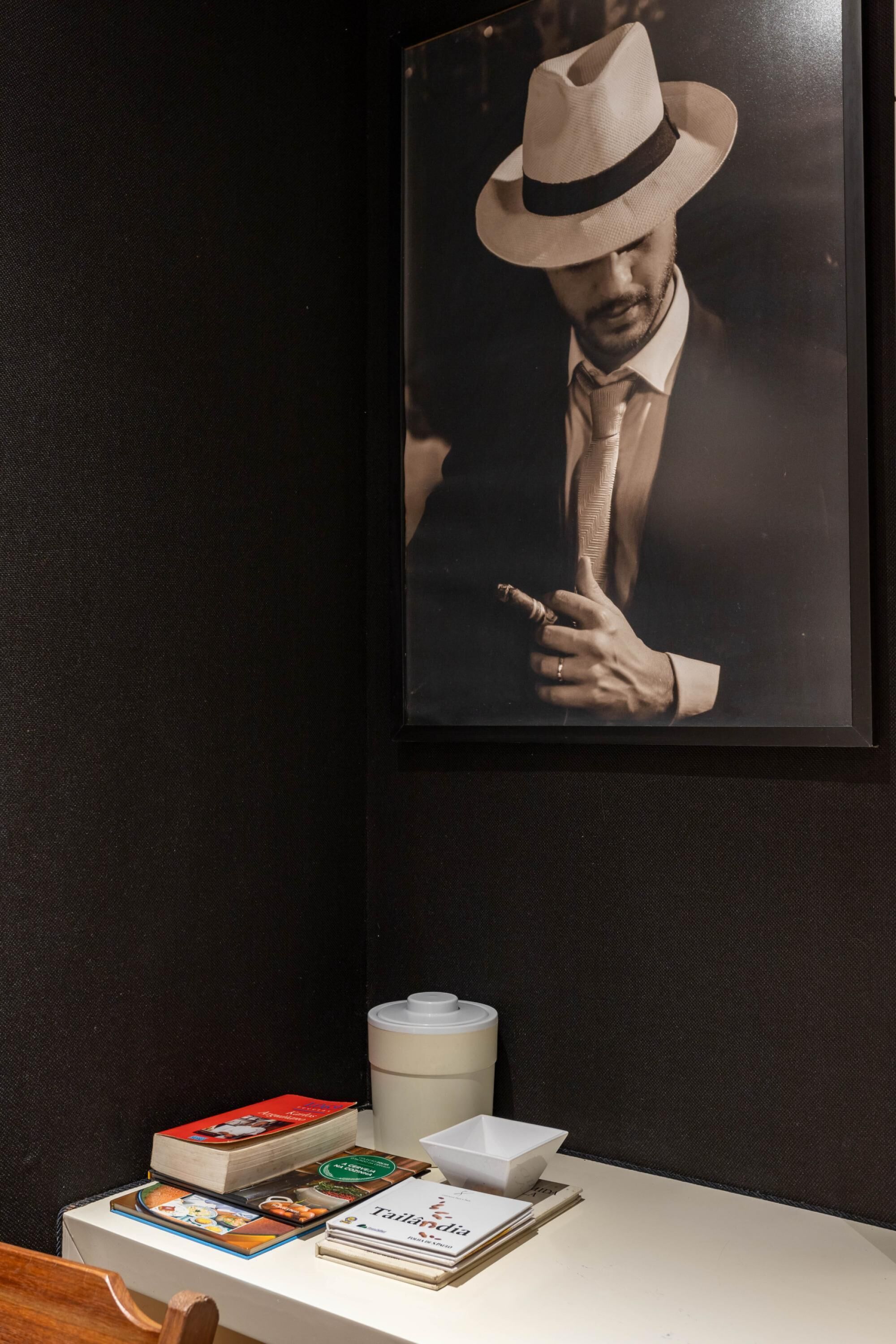 A framed photograph of a man in a hat and suit, a stack of books, and a white container on a table against a dark wall.