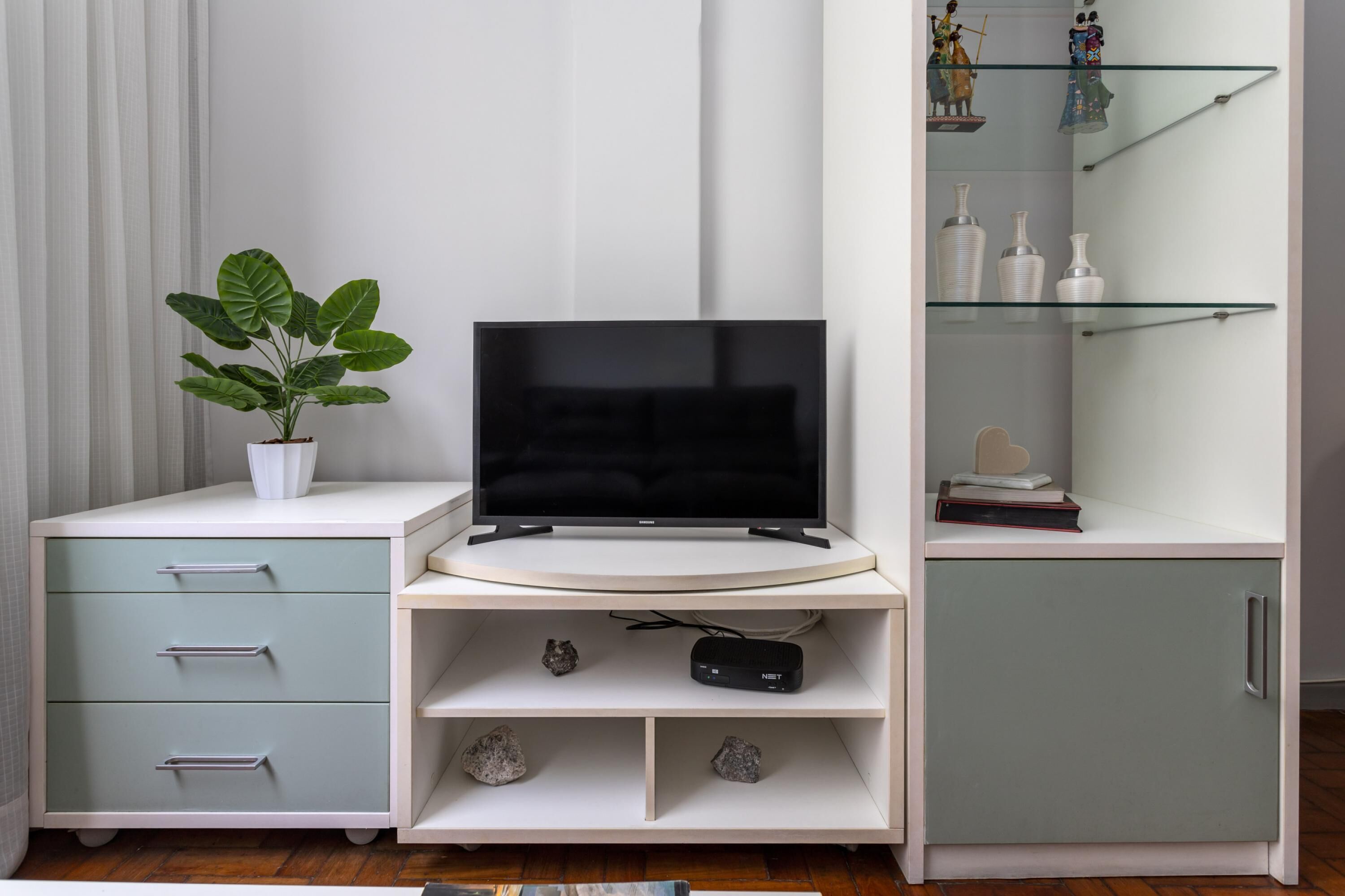 A modern living room with a flat-screen TV on a curved white entertainment unit, a potted plant, and a glass display cabinet with decorative items.