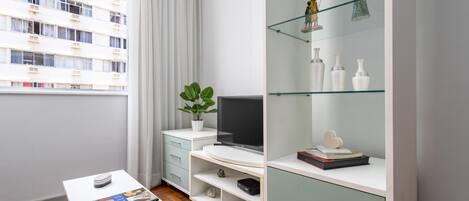 Un salon moderne doté d’un téléviseur à écran plat, d’une table basse blanche sur laquelle repose un magazine, d’une plante en pot et d’une vitrine en verre contenant des objets décoratifs.
