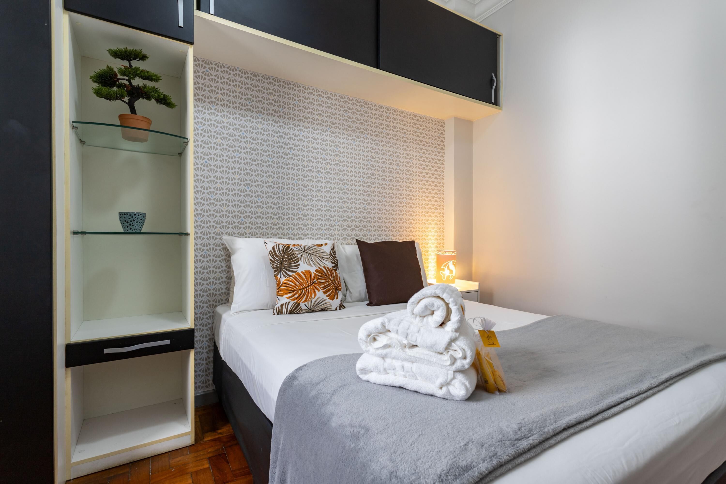 A neatly made bed with a grey blanket and a folded towel, a bedside table with a lamp, and a shelving unit with a potted plant and decorative items.