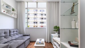 A modern living room with a grey sofa, a white coffee table, and a view of a multi-story building through a large window. - Pleasant in Copa Ideal for Friends Sc602 (Rio de Janeiro)