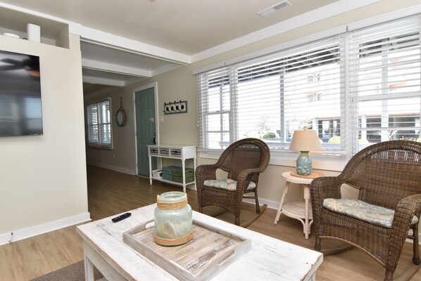 A cozy living room with wicker chairs, a small table with a lamp, and a television on the wall.
