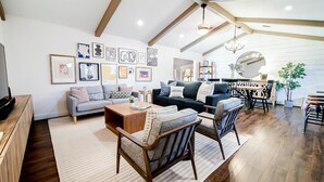 Un salon spacieux avec un plafond haut, des poutres apparentes, un coin repas avec une table ronde et des chaises, et un coin salon confortable avec un canapé et des fauteuils.