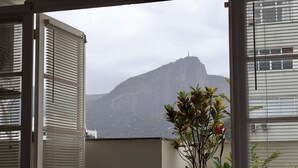 Vista a través de una ventana con persianas, una planta y una montaña al fondo.
