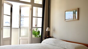 Una habitación de hotel con cama, ventana con cortinas, lámpara y un cuadro en la pared.