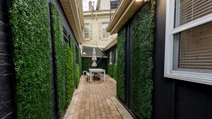 Une ruelle étroite avec un chemin pavé, bordée de murs recouverts de verdure, qui mène à un espace aménagé avec une table et des chaises.