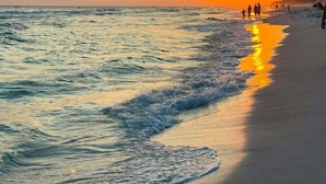 Een strand bij zonsondergang, met golven, mensen die wandelen en gebouwen op de achtergrond.