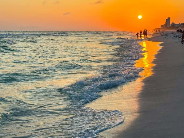 Una playa con olas al atardecer, gente caminando y edificios al fondo.