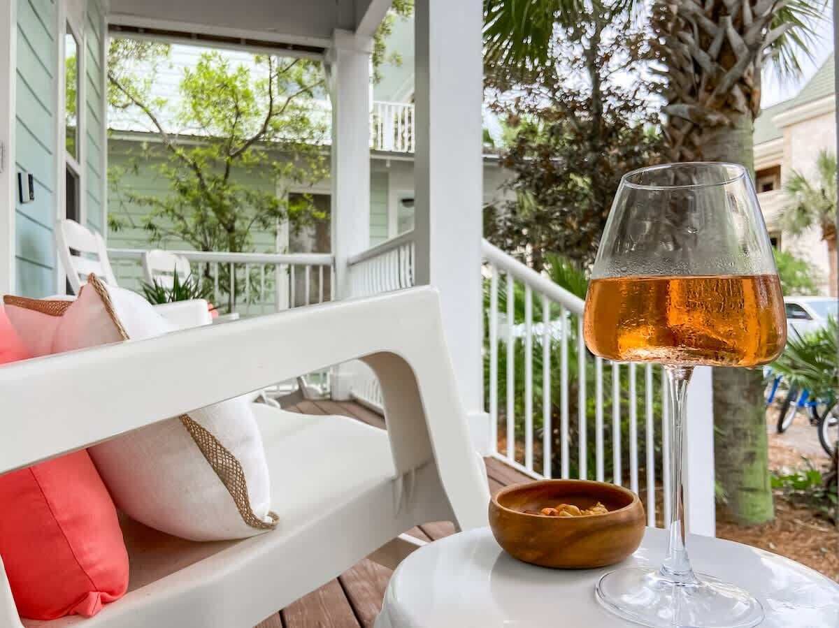 A white porch with a glass table, a white bench, and a glass of amber liquid.