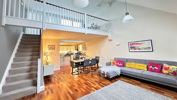 Un'ampia zona giorno con pavimento in legno, un divano grigio con cuscini colorati, un tavolo da pranzo con sedie e una scala che conduce a un piano superiore.