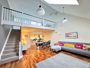 Un espacio habitable amplio con piso de madera, un sofá gris con cojines coloridos, una mesa de comedor con sillas y una escalera que sube a un nivel superior.