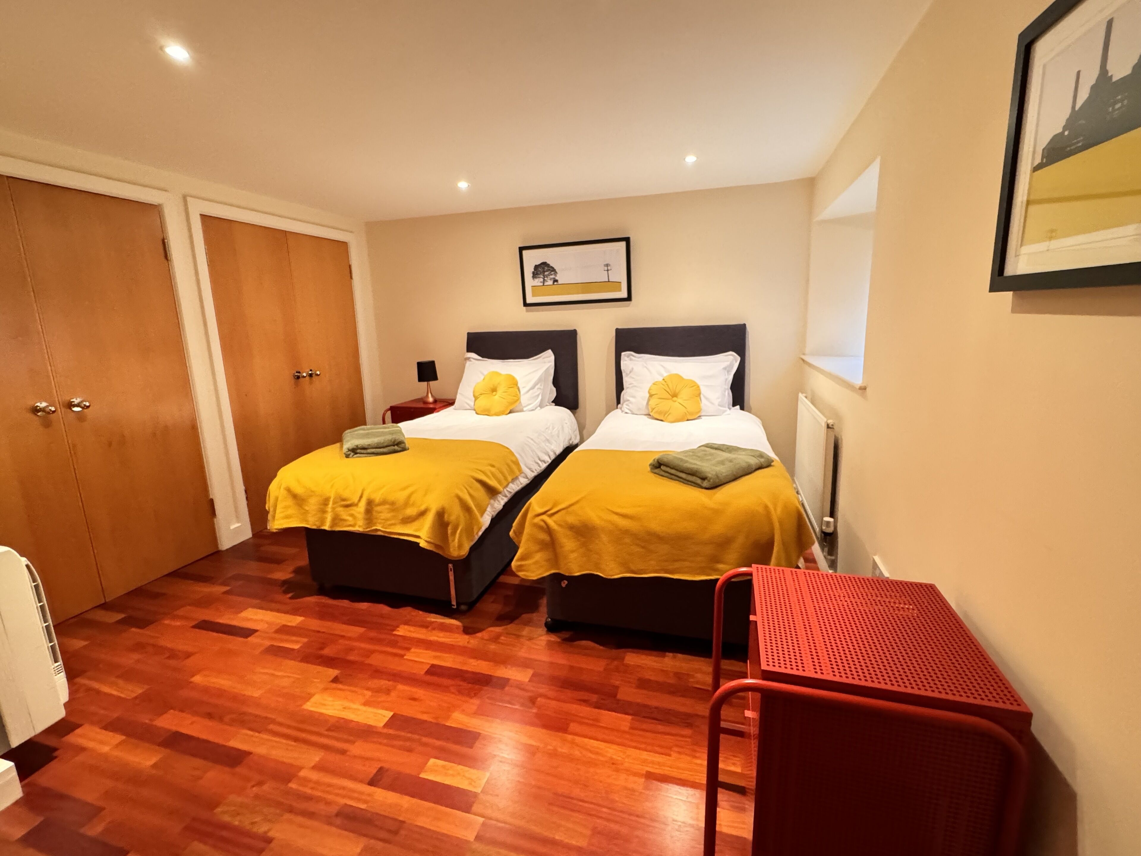 A room with two beds, wooden flooring, a red chair, and a framed picture on the wall.
