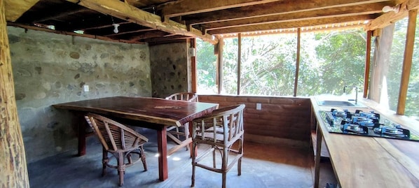 A rustic wooden dining area with a large table and chairs, a stone wall, and a window with a view of trees.