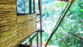 A rustic wooden structure with a red roof, bamboo walls, and a window overlooking a forested area.