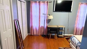 Une pièce avec un plancher en bois, un bureau avec une chaise, un téléviseur fixé au mur et un lit recouvert d’une couvre-lit à motifs.