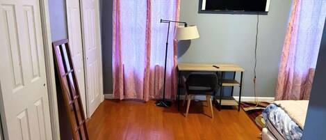 Una habitación con piso de madera, un escritorio con silla, un televisor montado en la pared y una cama con colcha estampada.