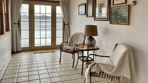 Et værelse med udsigt over en sø, en dør der fører ud, en stol, et bord med en lampe og flere billeder i rammer på væggen.
