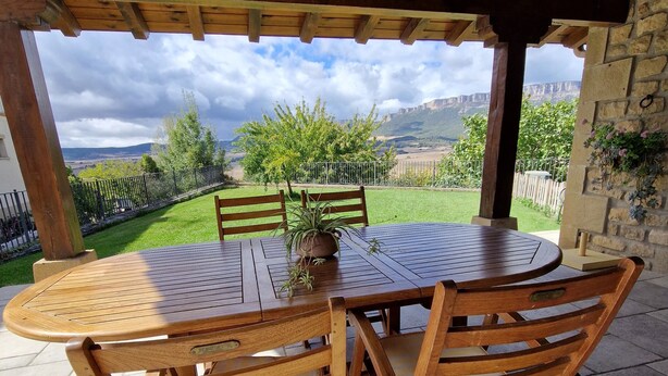 A wooden outdoor patio with a round table and chairs, a stone wall with greenery, and a view of a hilly landscape.