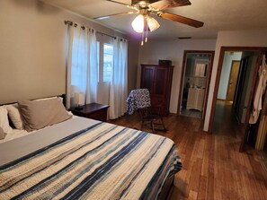 Quarto com cama, piso de madeira, ventoinha no teto, guarda-roupa e banheiro visível por uma porta aberta.