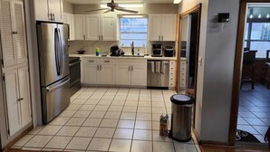 A compact kitchen with white cabinets, a stainless steel refrigerator, a ceiling fan, and a window providing natural light.