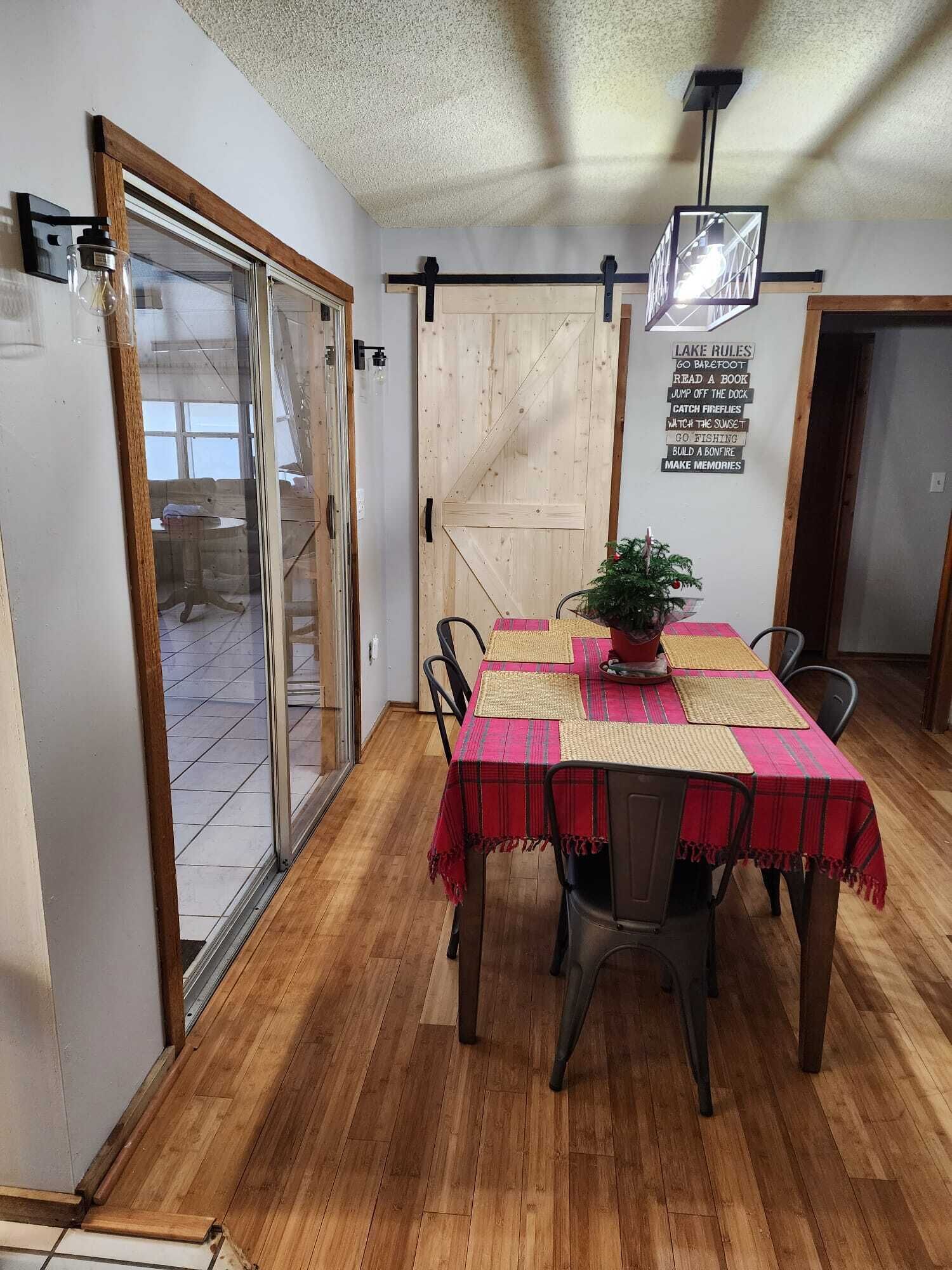 Een eethoek met een houten tafel, stoelen en een rode tafelkleed. Er is een schuifdeur van glas die leidt naar een balkon, en een bord op de muur met activiteiten.