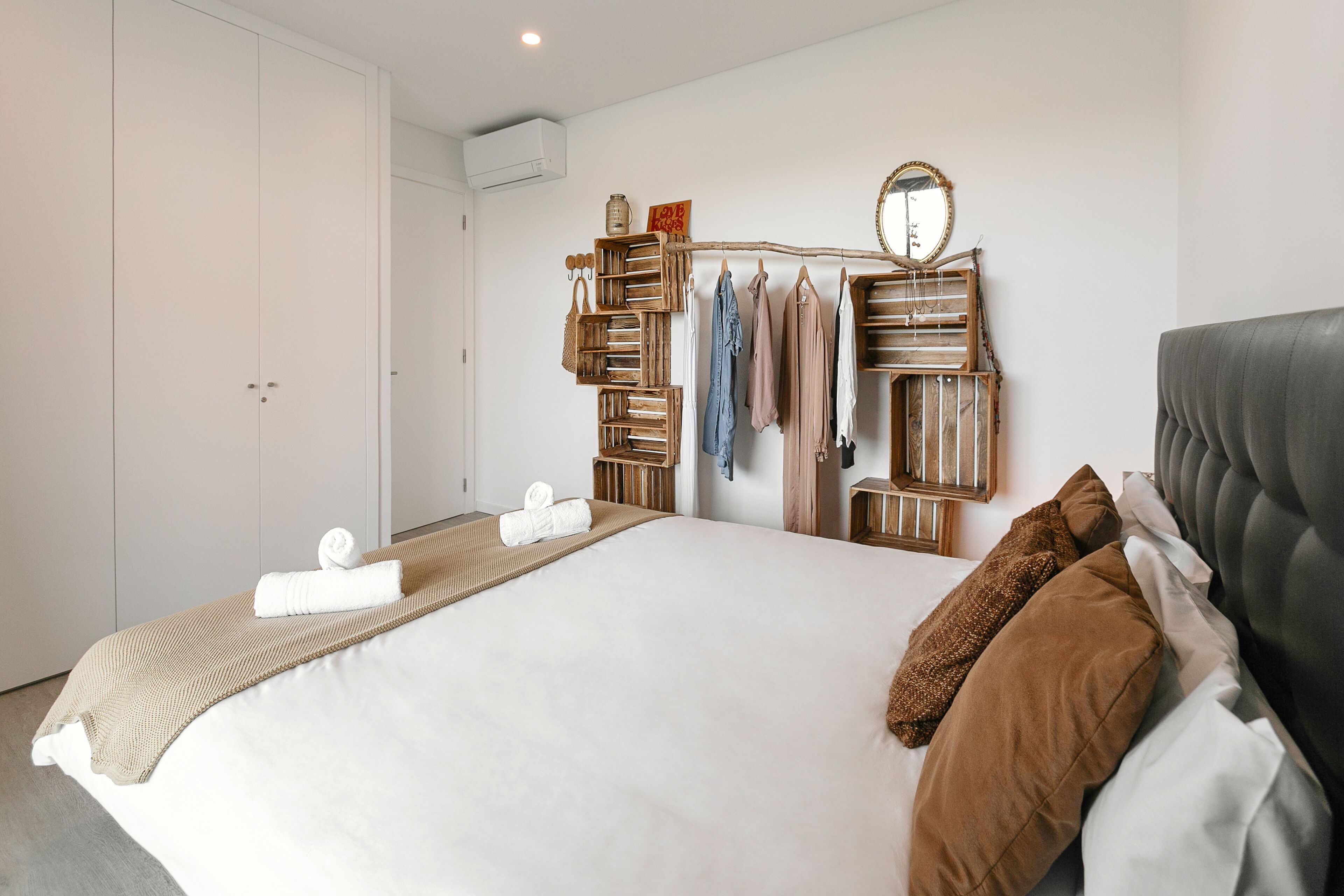 A neatly arranged bedroom with a bed, a wardrobe, a mirror, and a hanging rack with clothes and bags.