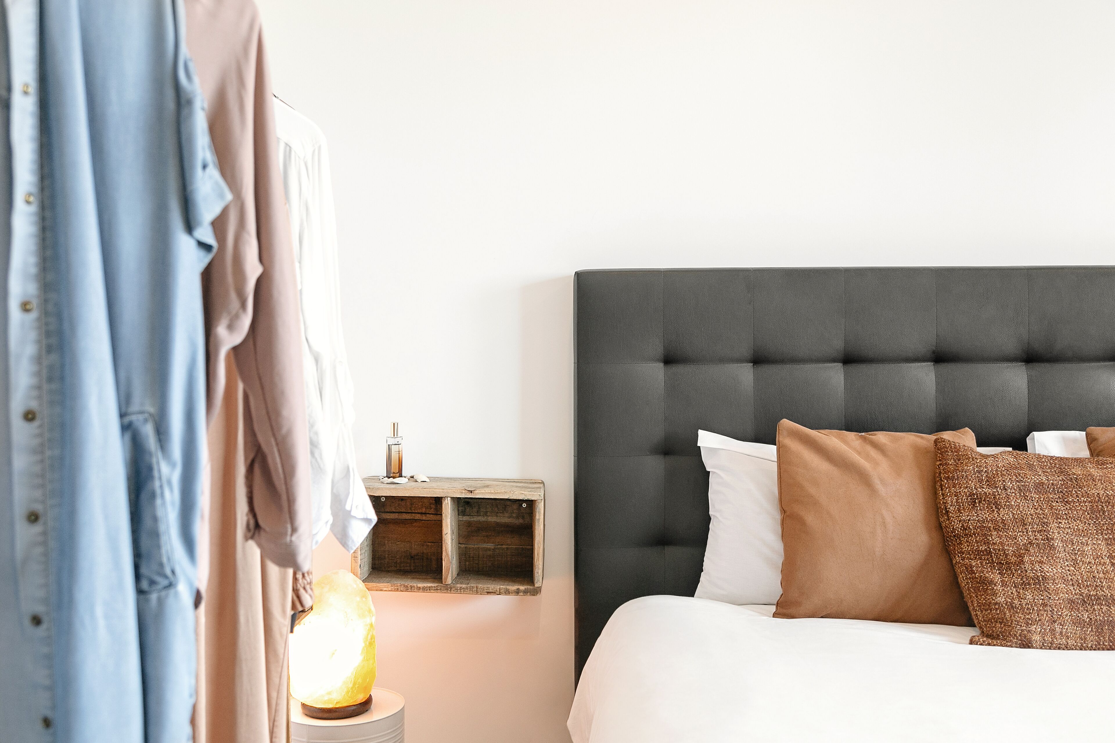 Quarto com cabeceira estofada, mesa de cabeceira com abajur e guarda-roupa com roupas penduradas.
