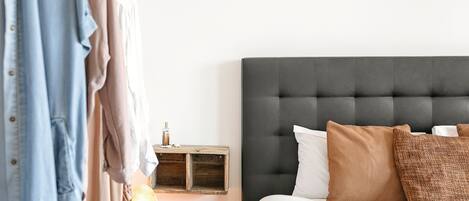 Quarto com cabeceira estofada, mesa de cabeceira com abajur e guarda-roupa com roupas penduradas.