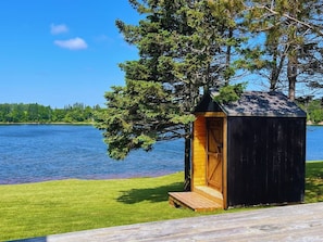 A small wooden structure with a door and steps, located near a body of water and surrounded by grass and trees. - Waterfront with access, 3BR, Dock, Sauna & Hot Tub (Alberton)