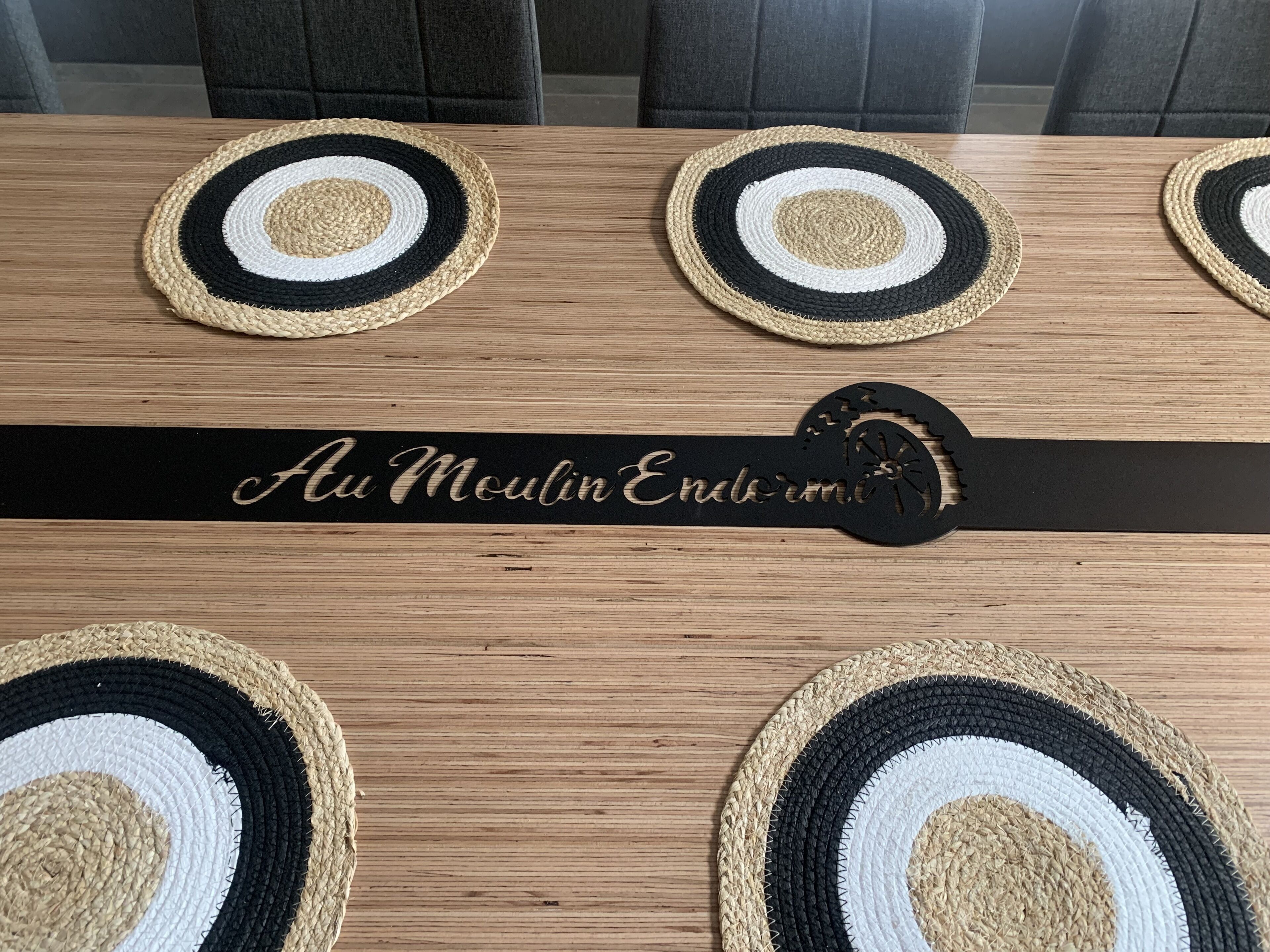 Une table en bois surmontée de quatre sets de table ronds en fibres tressées, de couleur noire, blanche et beige. Une baguette noire portant l’inscription « Au Moulin Endormé » est placée sur la table.