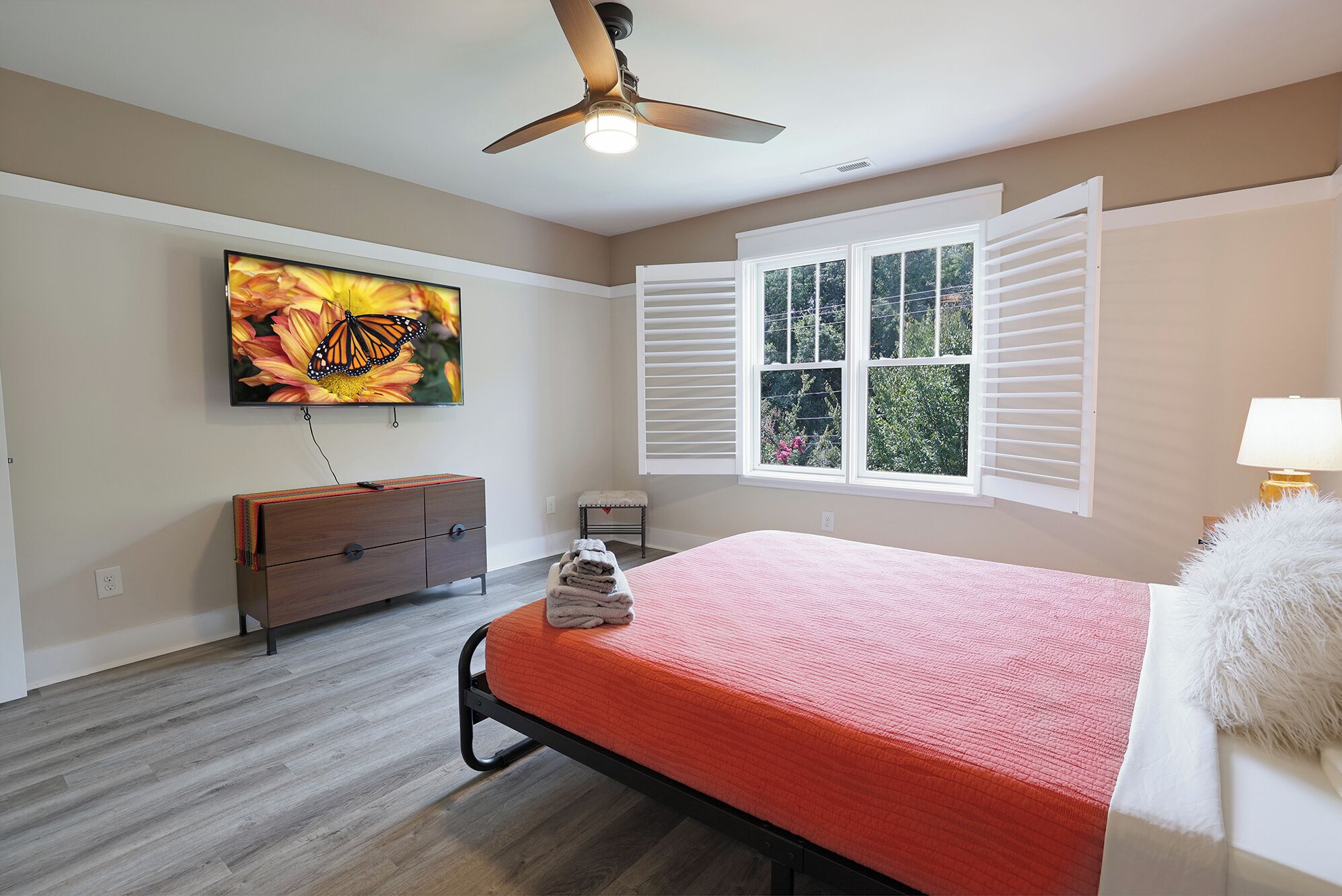 Quarto com cama, cómoda, ventoinha no teto, janela com persianas e televisão na parede.