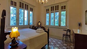Une chambre à coucher comprenant un lit, un bureau, une chaise, une lampe, un miroir et de grandes fenêtres donnant sur la forêt.