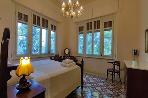 A bedroom with a bed, a desk, a chair, a lamp, a mirror, and large windows with a view of trees.