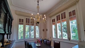 Une salle à manger avec une grande table, des chaises, un lustre et de grandes fenêtres équipées de volets.