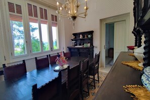 A dining area with a large table, chairs, a chandelier, and a view of greenery outside.
