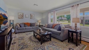 A living room with two sofas, a coffee table, and a TV. There are framed pictures on the wall and a large window with curtains.