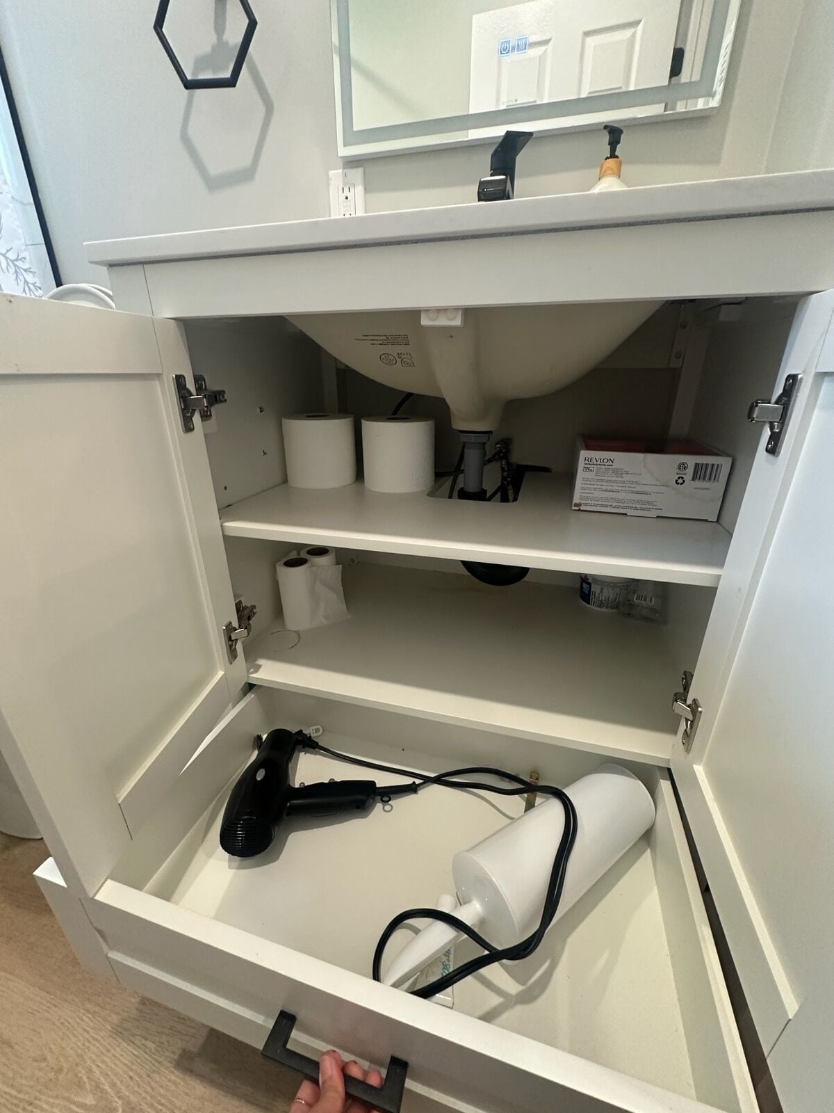 A white cabinet with three shelves, containing toilet paper, a box, and a hairdryer.