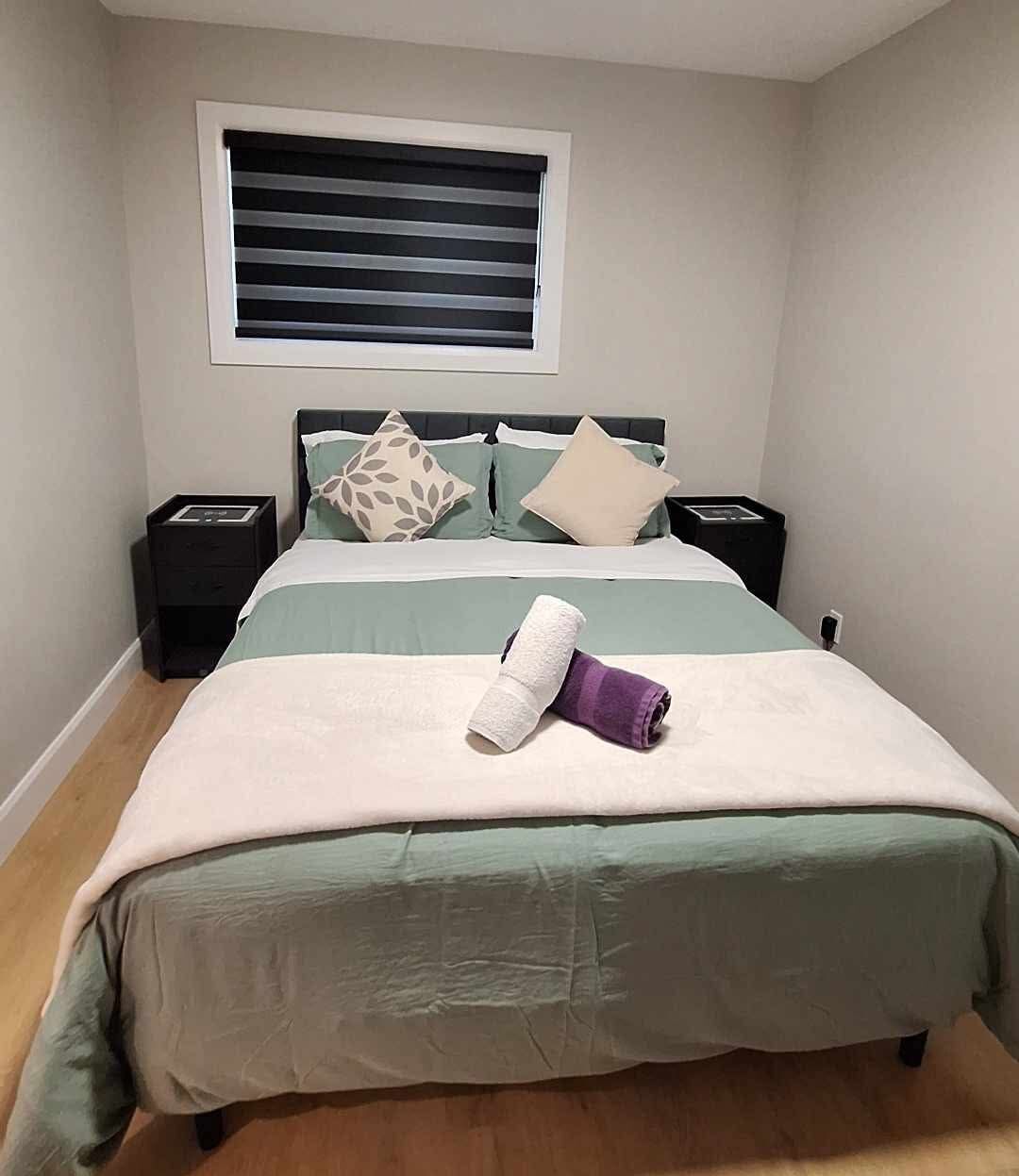 A neatly made bed with a green and white bedspread, two pillows, a rolled towel, and a bedside table with a lamp.