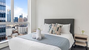 Une chambre d’hôtel moderne avec un grand lit, une table de chevet, une lampe et une vue sur le paysage urbain, visible à travers une grande fenêtre.