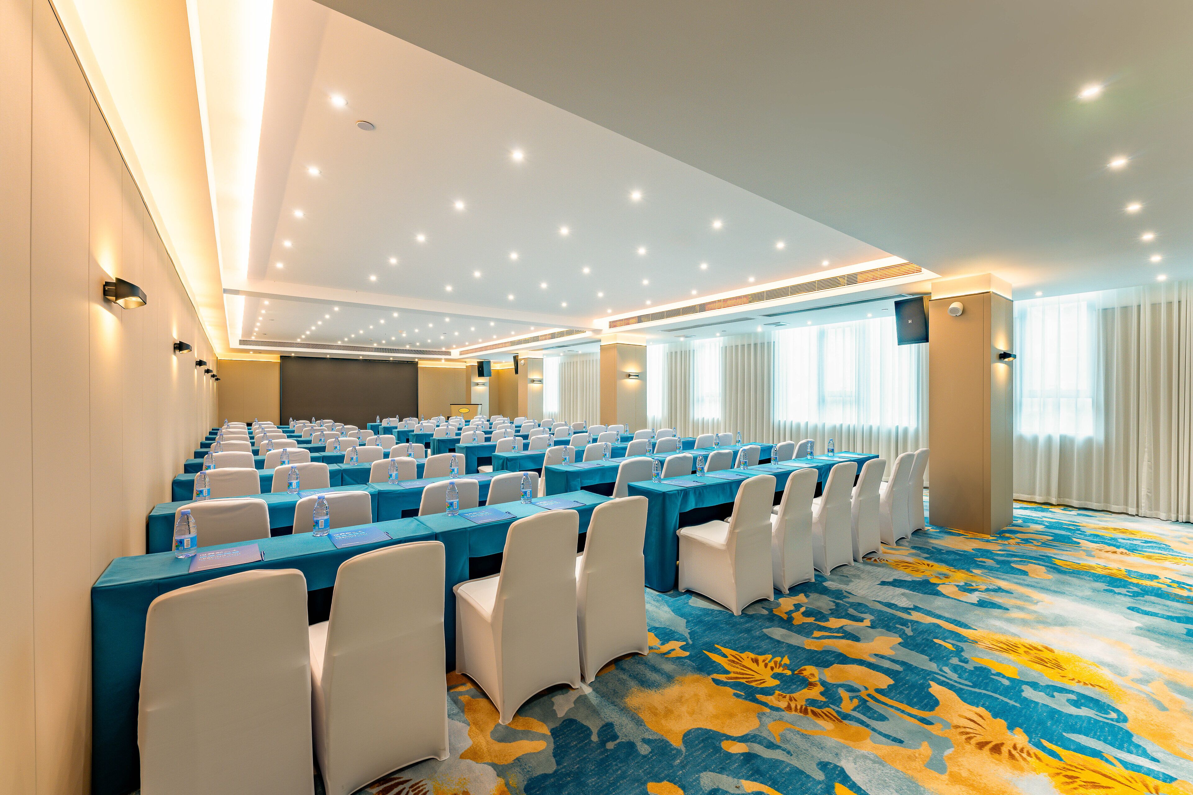 A conference room with rows of tables, chairs, and water bottles, featuring a patterned carpet and ceiling lights.