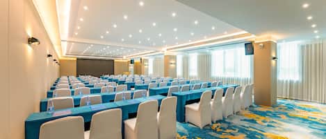 A conference room with rows of tables, chairs, and water bottles, featuring a patterned carpet and ceiling lights.