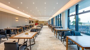 Un vaste espace intérieur aménagé en salle à manger, avec de nombreuses tables et chaises, une cuisine à l’arrière-plan et de grandes fenêtres offrant une vue dégagée.