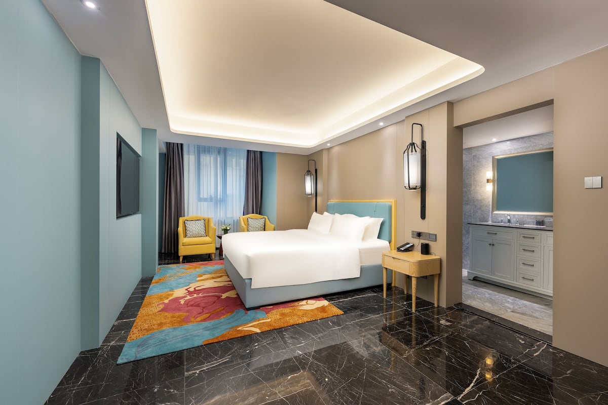 A modern hotel room with a large bed, a small desk, a nightstand, a bathroom with a mirror, and a kitchen area.