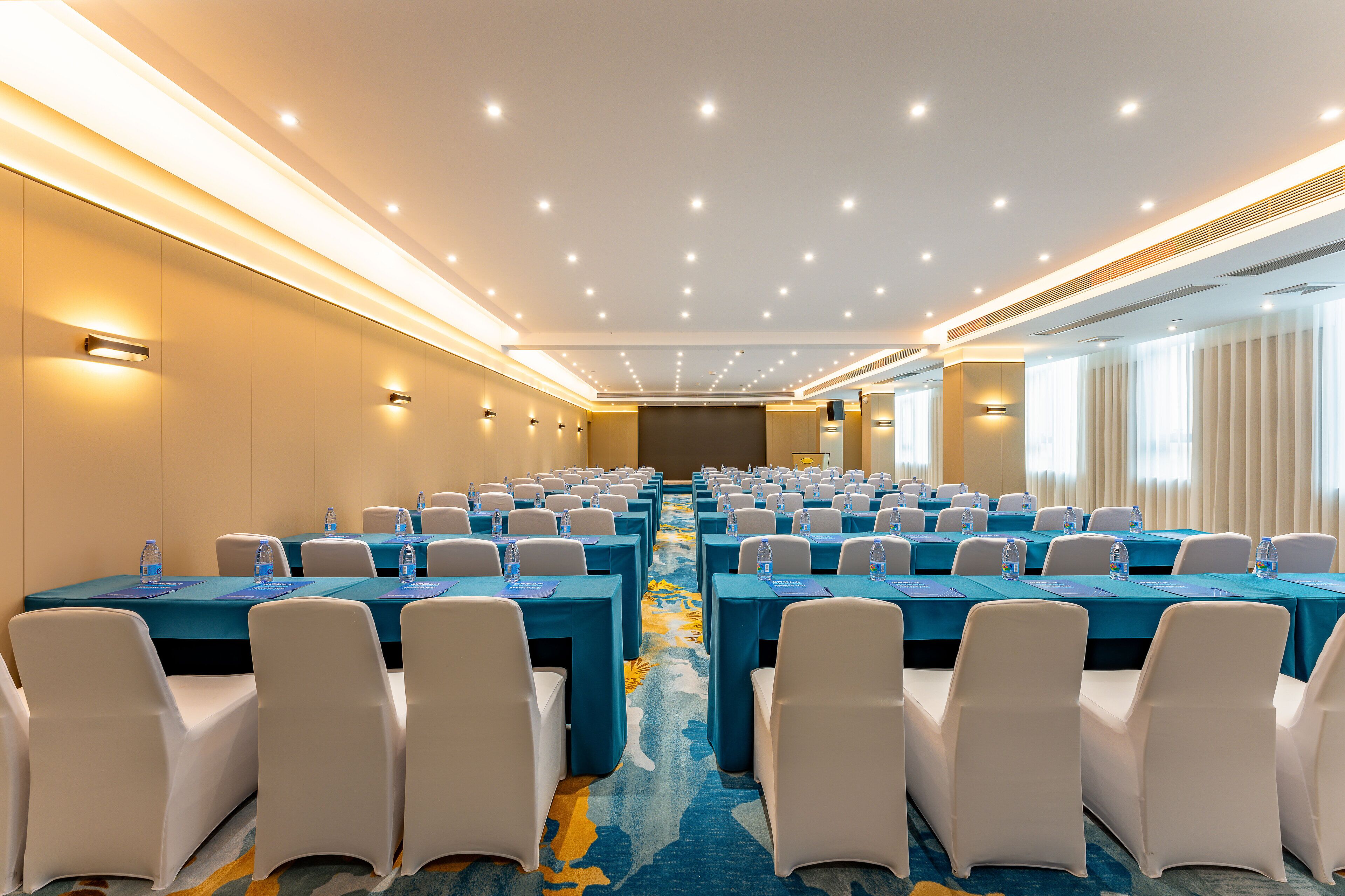A conference room with rows of tables, chairs, and water bottles, featuring a carpeted floor and ceiling lights.
