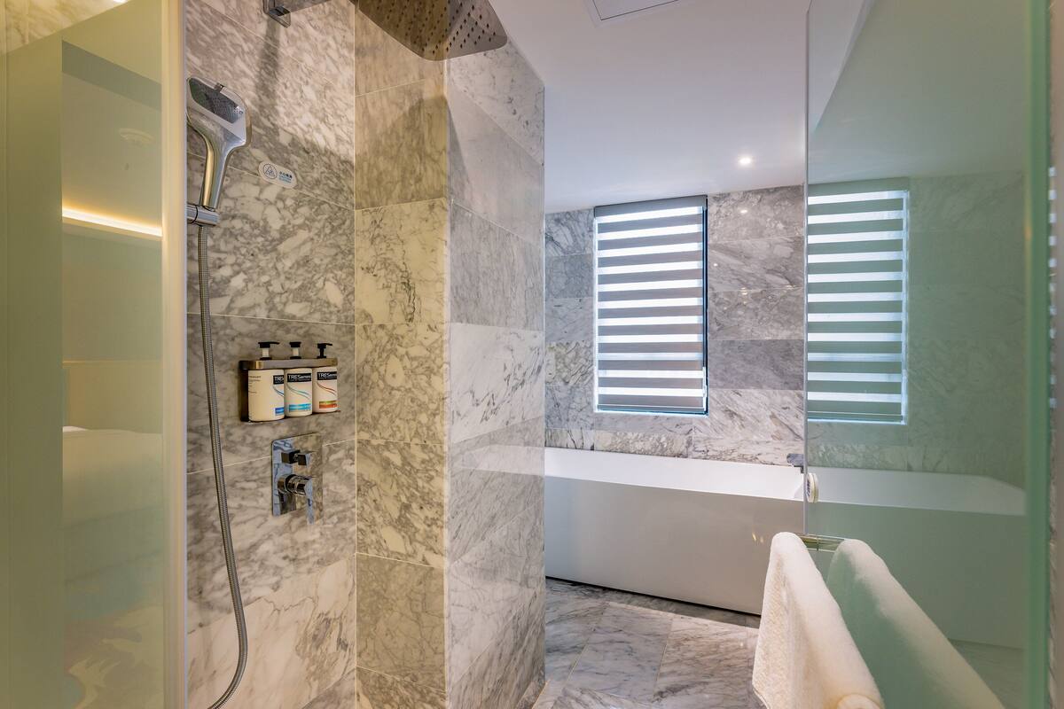A modern bathroom with a marble floor, a shower with a handheld showerhead, a bathtub, and a window with blinds.